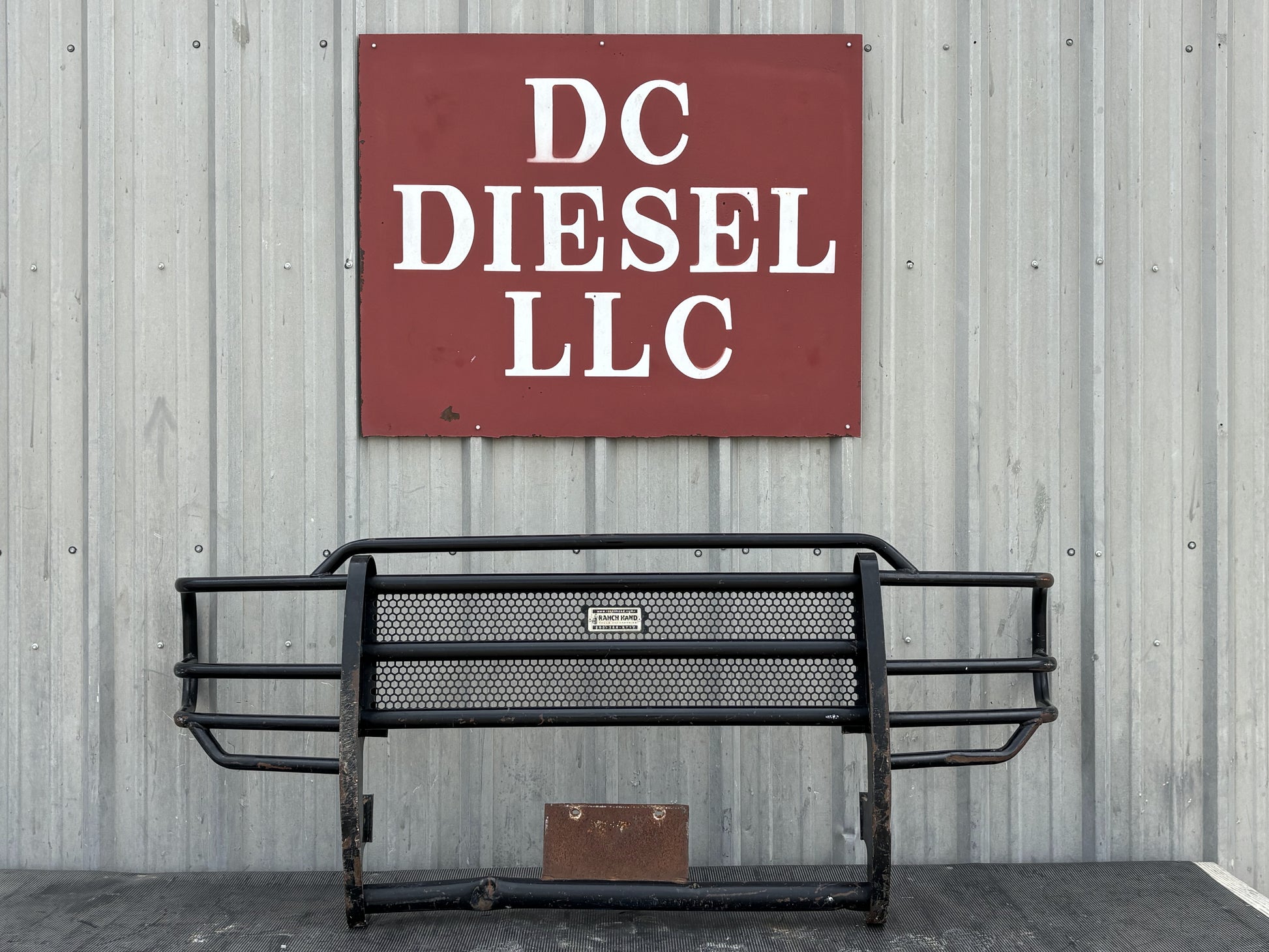 99-04 Ford F250 F350 Front Ranch Hand Grill Brush Guard in used condition with light wear and a bent lower pipe, displayed against a corrugated metal wall beneath a DC Diesel LLC sign, compatible with 1999-2004 Ford Super Duty models including 6.0L Powerstroke engines.