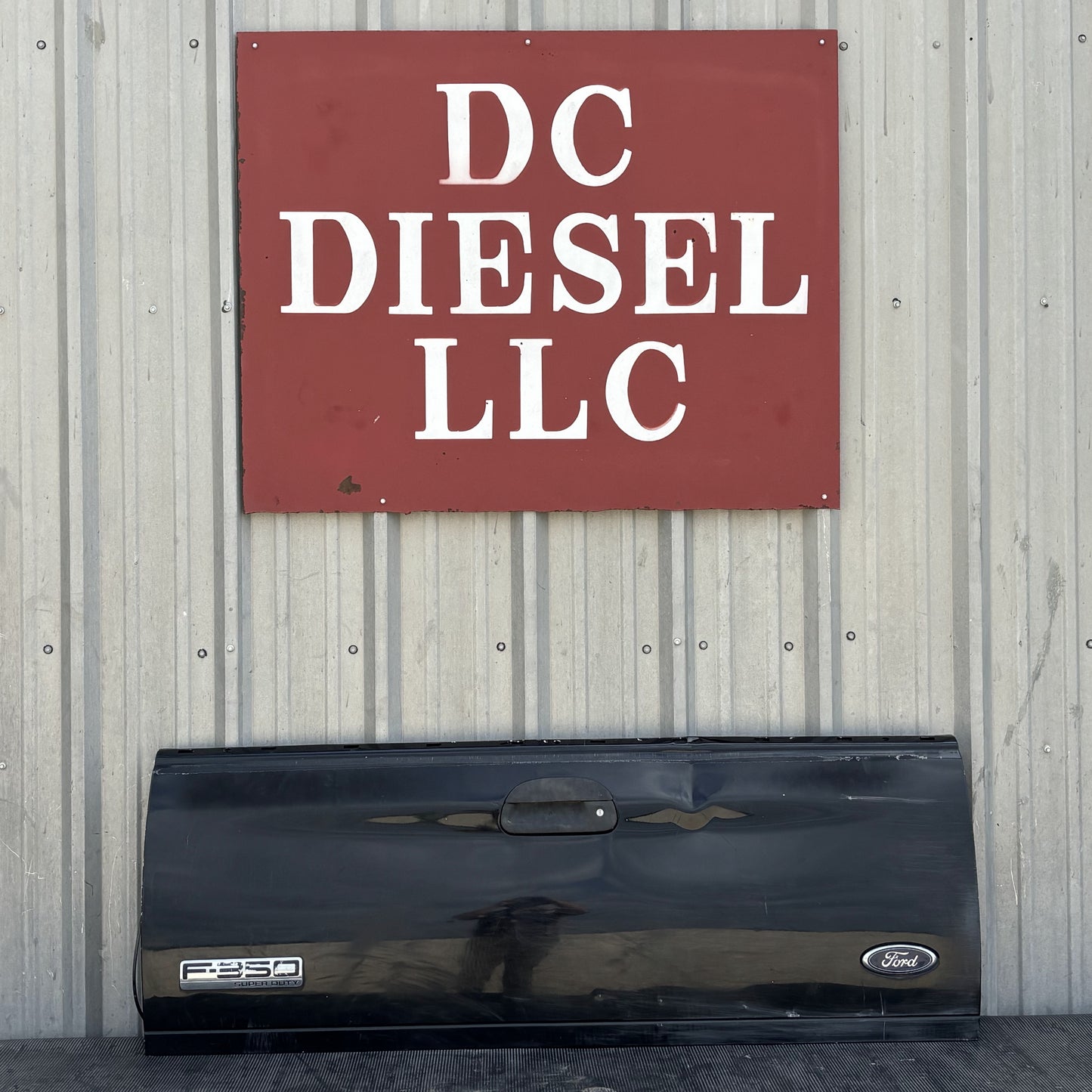 99-07 Ford F250 F350 Tailgate UD Black with Ford and F250 badges, showing light wear and dent, displayed against a corrugated metal wall beneath a DC Diesel LLC sign, suitable for 6.0L Powerstroke and Super Duty Ford trucks.