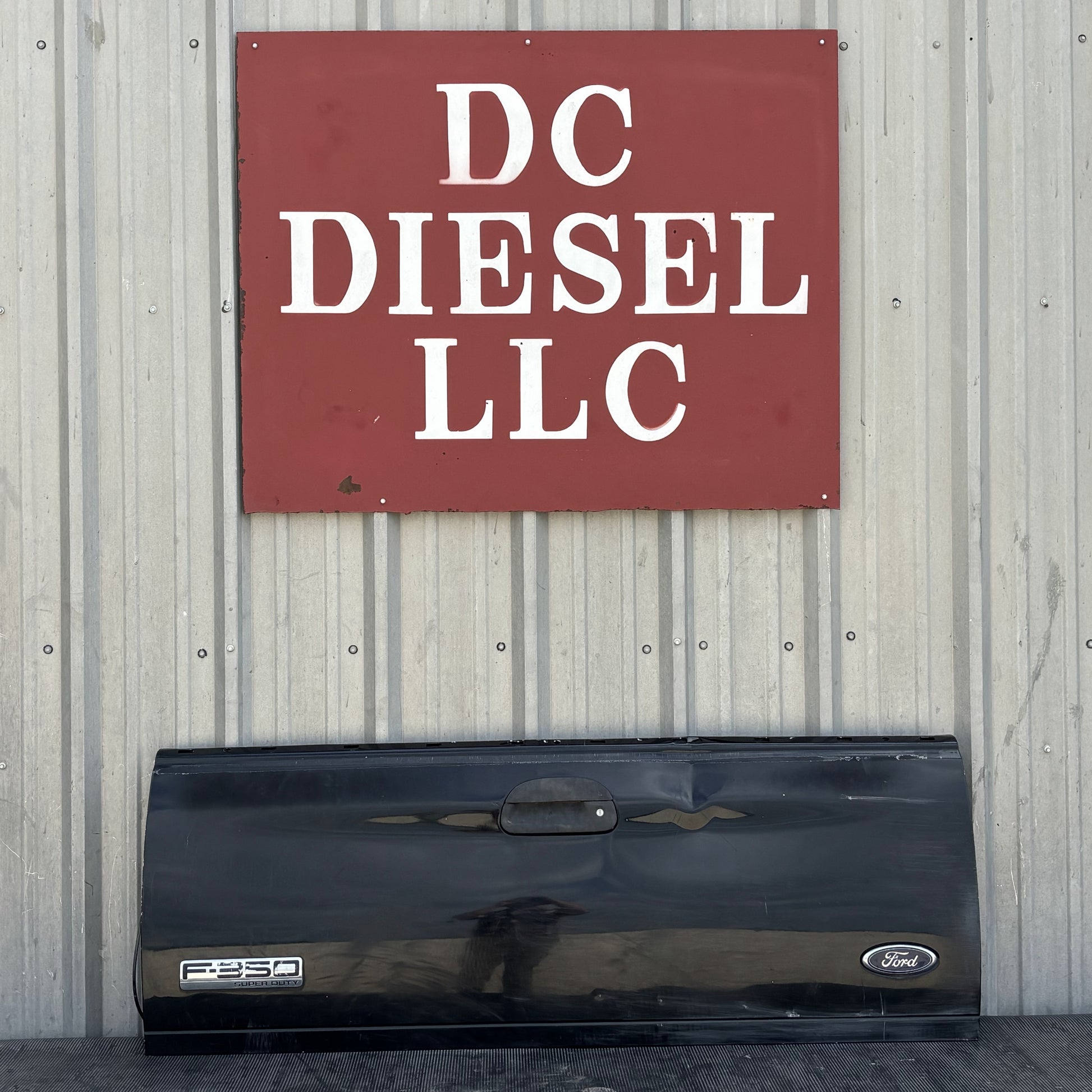 99-07 Ford F250 F350 Tailgate UD Black with Ford and F250 badges, showing light wear and dent, displayed against a corrugated metal wall beneath a DC Diesel LLC sign, suitable for 6.0L Powerstroke and Super Duty Ford trucks.