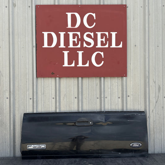 99-07 Ford F250 F350 Tailgate UD Black with Ford and F250 badges, showing light wear and dent, displayed against a corrugated metal wall beneath a DC Diesel LLC sign, suitable for 6.0L Powerstroke and Super Duty Ford trucks.