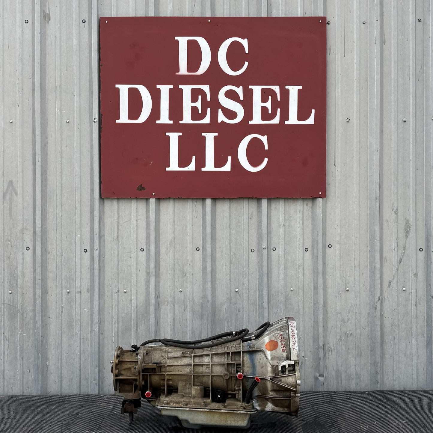 03-07 Ford F250 F350 Diesel 5R110 5R110W Automatic Transmission W/ PTO displayed in front of a metal wall with a DC Diesel LLC sign, showing the used transmission removed from a 2004 Ford F-350 Lariat 6.0L Powerstroke truck, suitable for Ford Super Duty models from 2003 to 2007.
